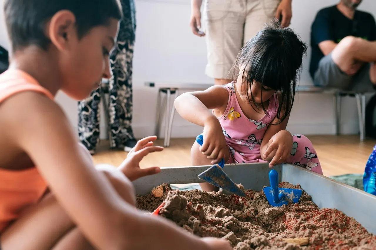 Young children playing and digging in a sand tray with toys whilst adults supervise nearby.