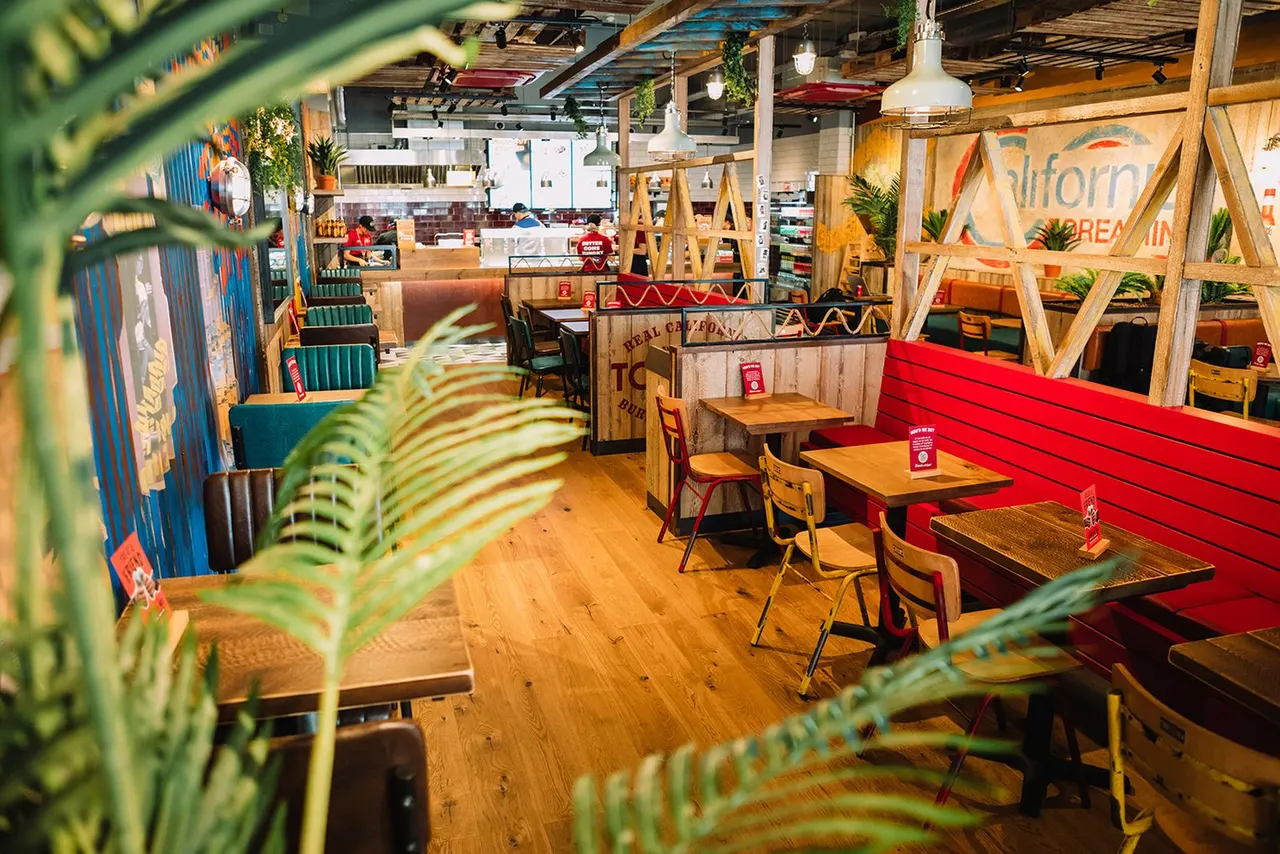 Colourful casual dining restaurant interior with wooden tables, chairs and decorative geometric screens, plants in foreground.