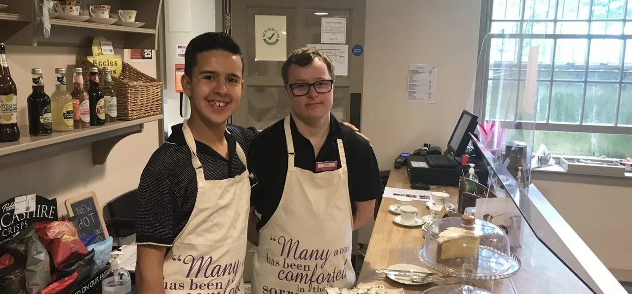 Two young staff members wearing aprons stand smiling behind a café counter with shelves of bottles and cakes.