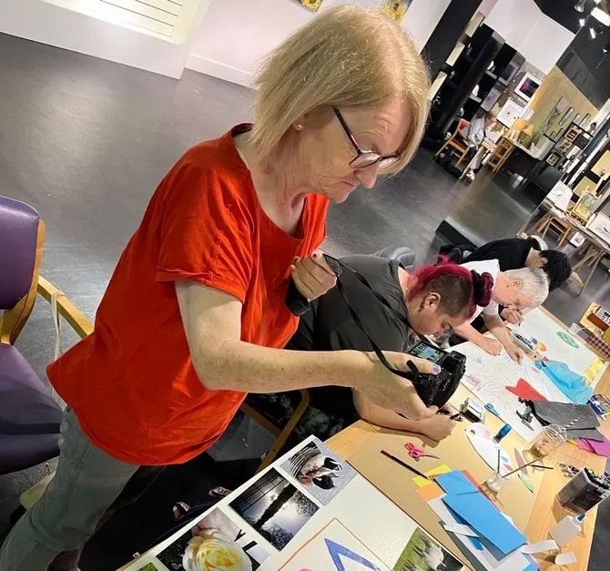 An adult in a red shirt leads a creative workshop, helping children with art materials and drawings at a table indoors.