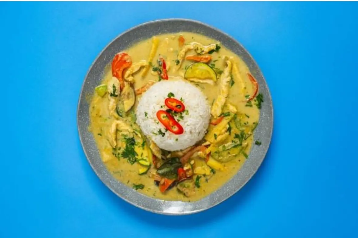 Curry dish with chicken, vegetables and white rice topped with sliced red chilli on a grey plate against blue background.