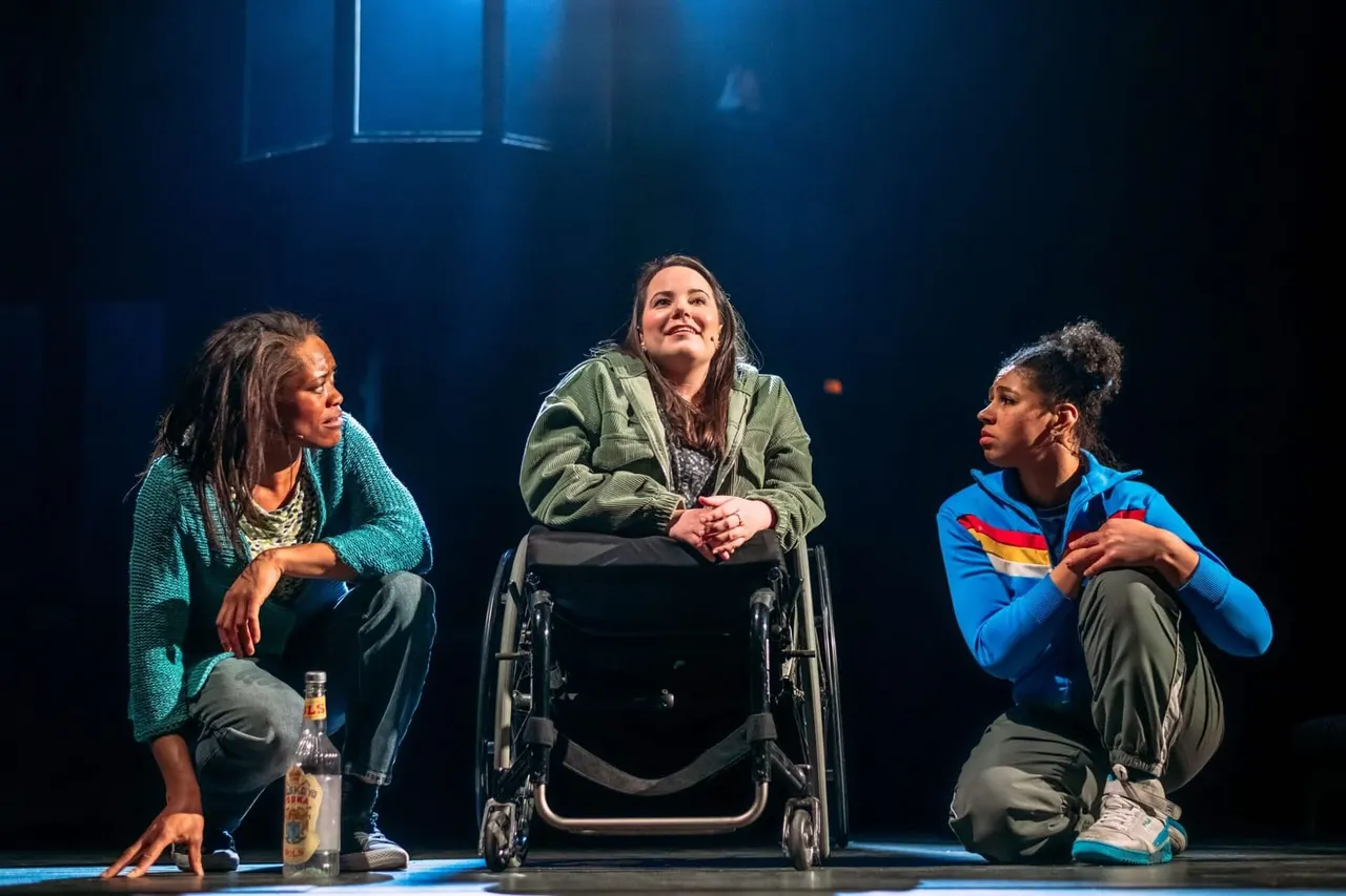 Three performers sit on stage during a theatrical scene, with the centre performer in a wheelchair.