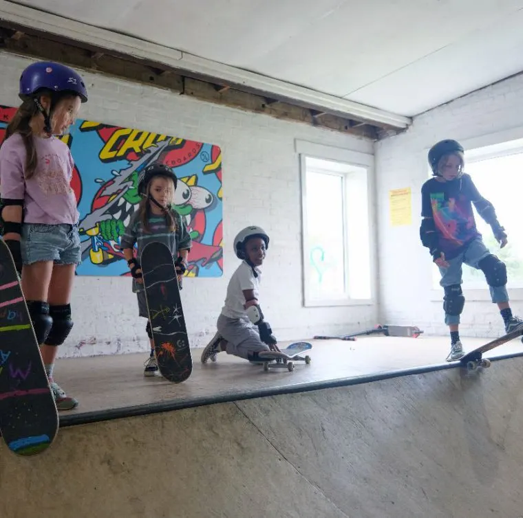 Children skateboarding indoors in a skate park with colourful wall art and protective gear.