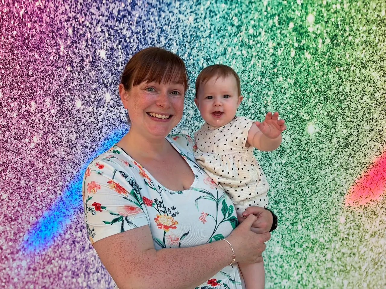 Woman holding a smiling toddler in front of a colourful rainbow glitter backdrop.
