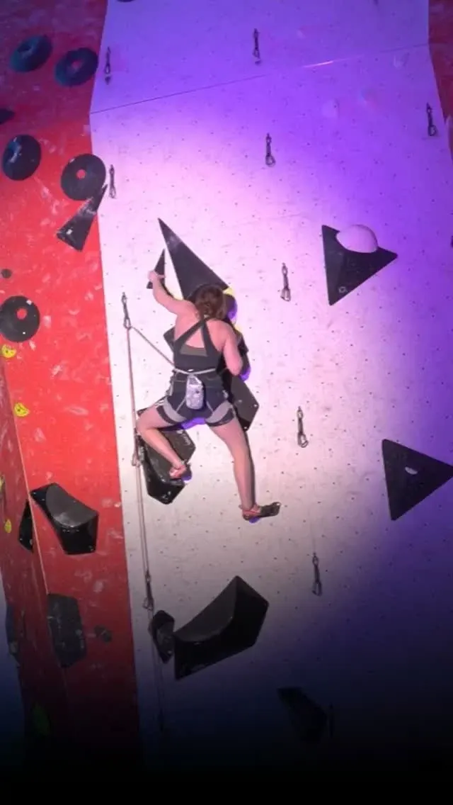 Person climbing an indoor rock wall with holds under purple and red lighting.