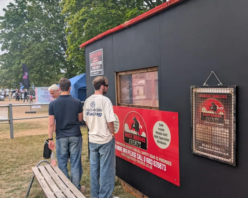 Two men standing outside a mobile rage room unit at an outdoor event, with trees and other visitors visible in the background.