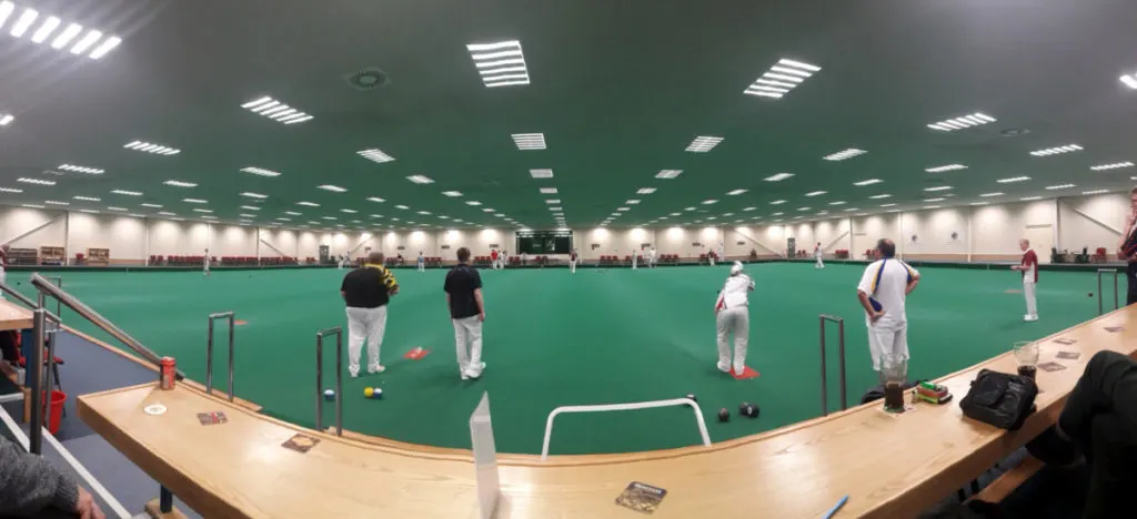 Players in white clothing playing bowls on a green indoor surface under bright lights.