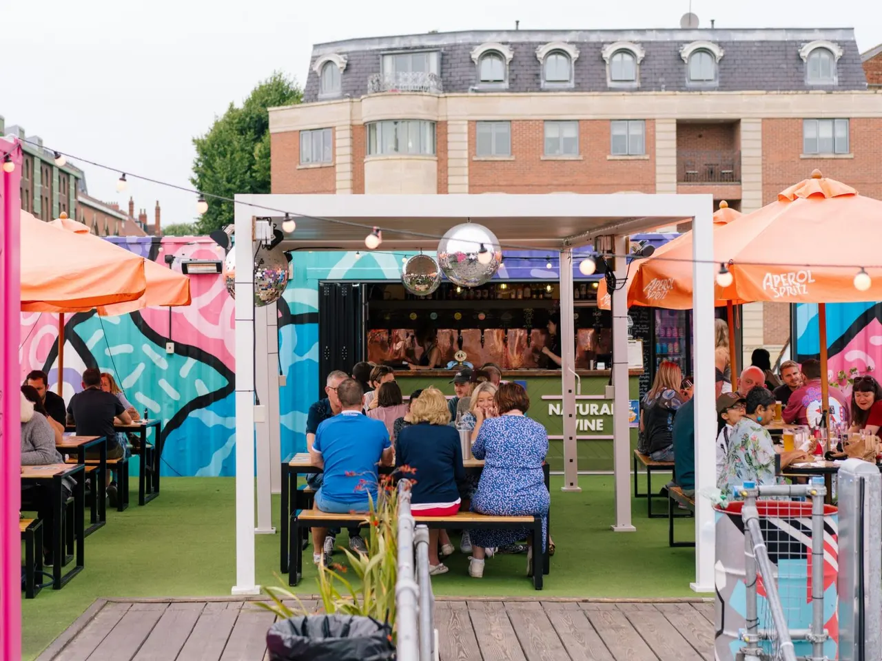 Outdoor event space with colourful umbrellas and covered seating areas where visitors socialise and dine at tables on artificial grass.