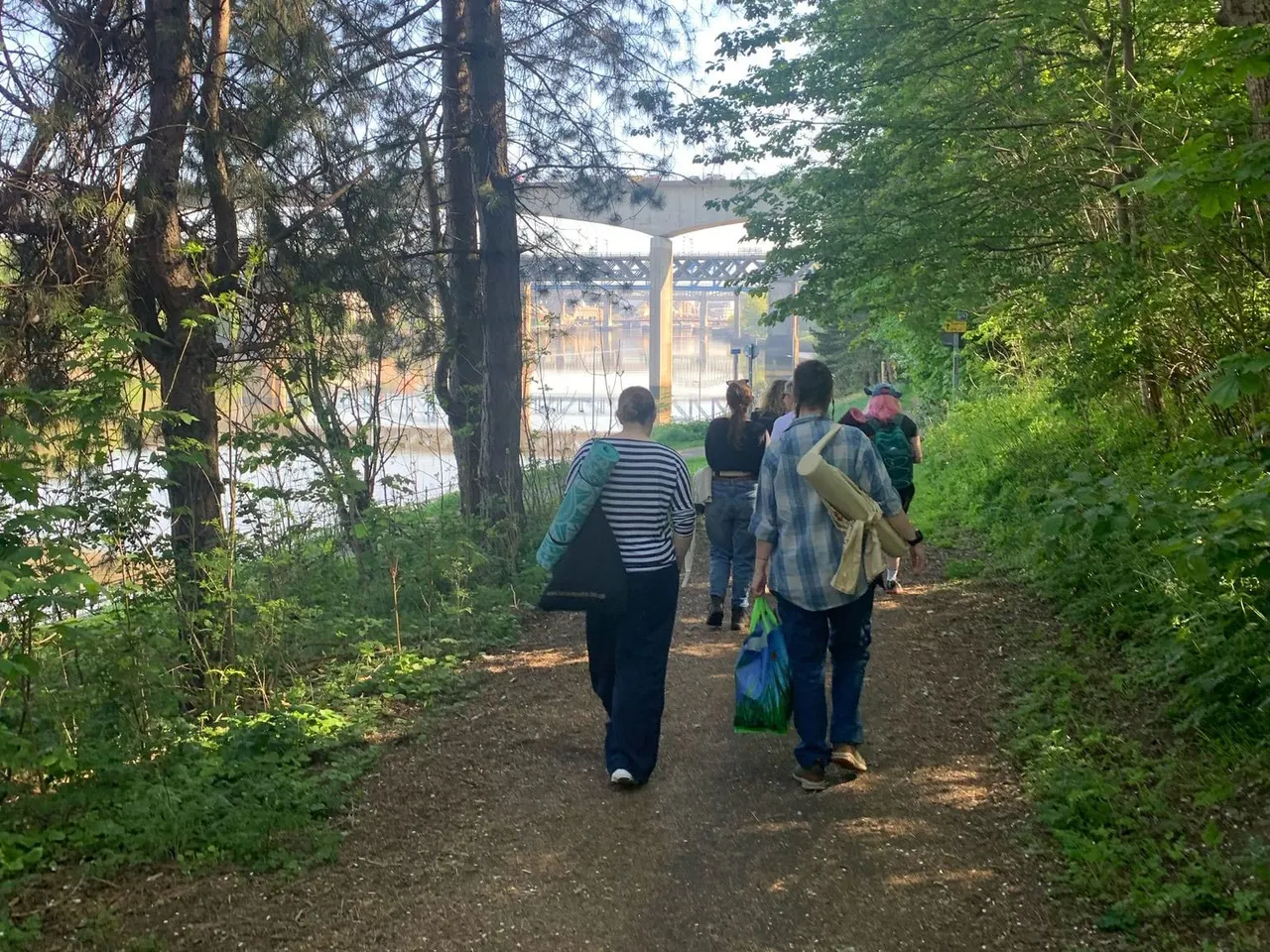 A group of visitors walk along a tree-lined path towards a waterside building surrounded by lush green vegetation.