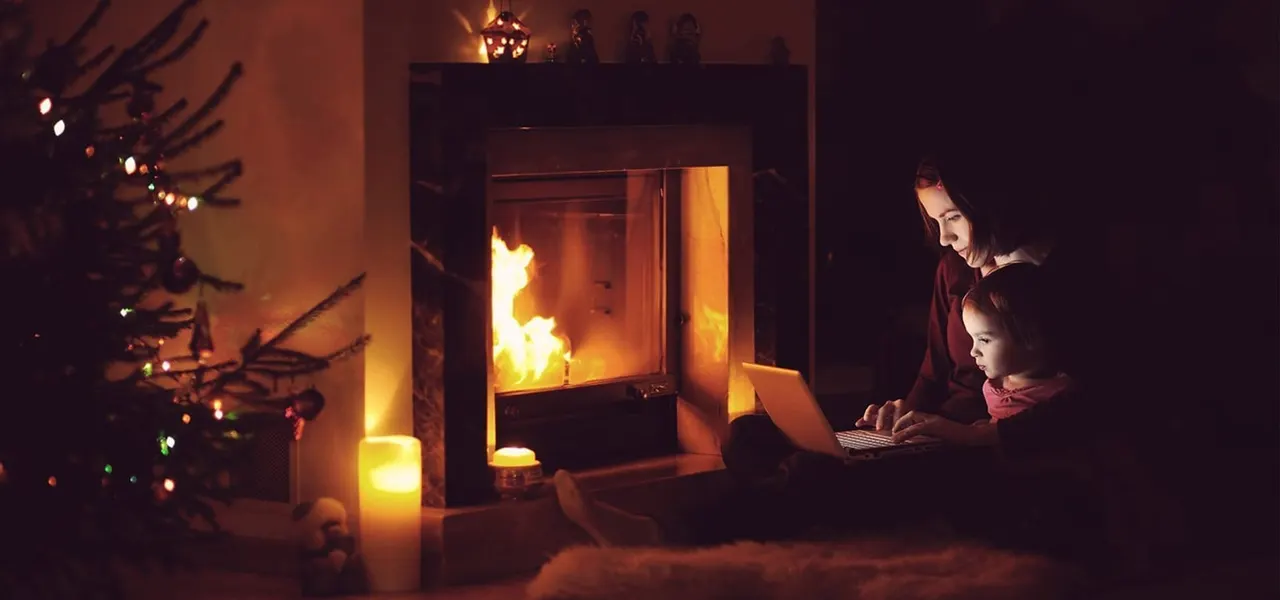 Woman and child sitting by a warm fireplace on a winter evening, with a decorated Christmas tree nearby.