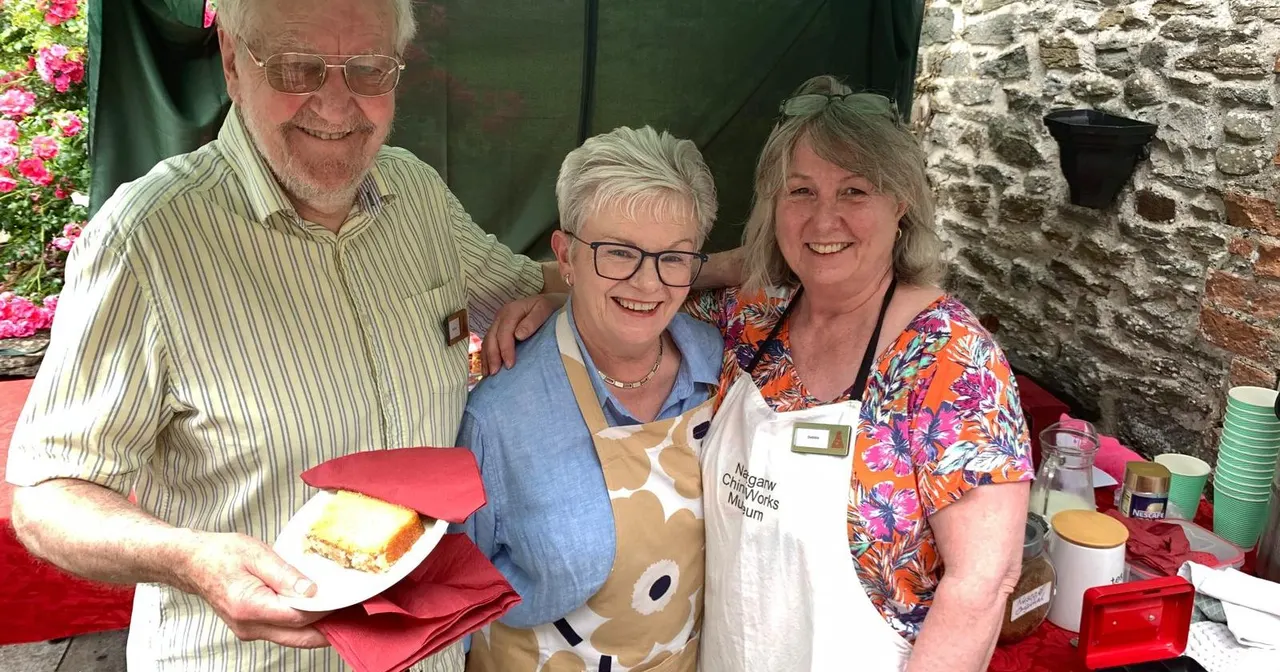 Three smiling adults at an outdoor event, with one holding a plate of food, standing in front of a stone building with pink flowers and a green canopy.
