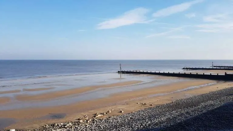 Sandy beach with pebbled shoreline, calm sea, and wooden pier extending into the water on a clear day.