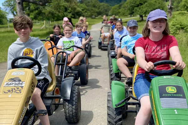 Children and teenagers driving toy tractors and vehicles along a tree-lined path during an outdoor activity at a farm park.