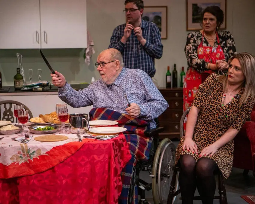 An elderly man in a wheelchair at a dining table gestures with a knife whilst other actors watch during a theatrical performance in a kitchen setting.