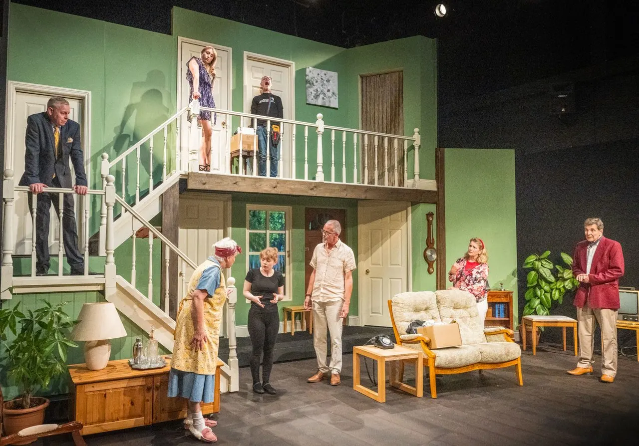 Actors perform a period comedy on a stylised two-level set with green walls, mid-century furniture, and a split staircase.