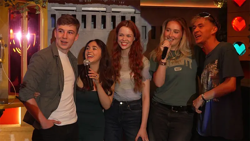 Five young people holding microphones singing together at a karaoke venue with neon heart decorations.