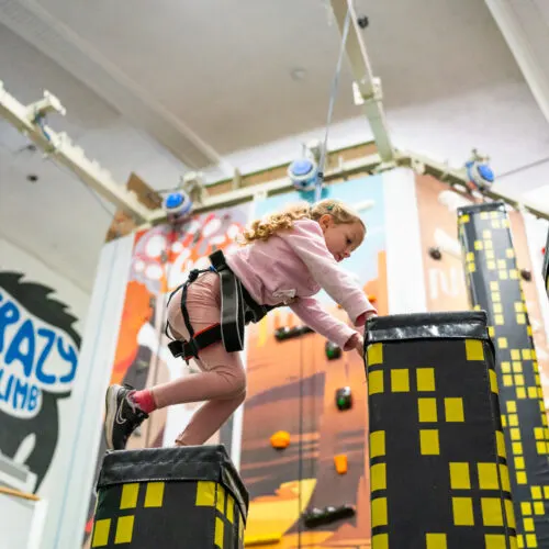 Child in harness climbing an indoor rock wall with illuminated building-themed obstacles.