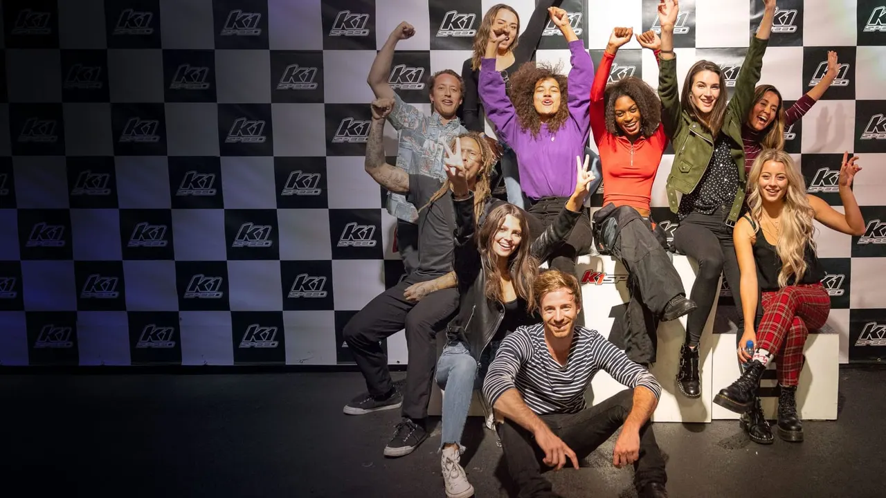 Group of adults and children posing together with raised arms in front of a chequered branded backdrop at an indoor racing venue.