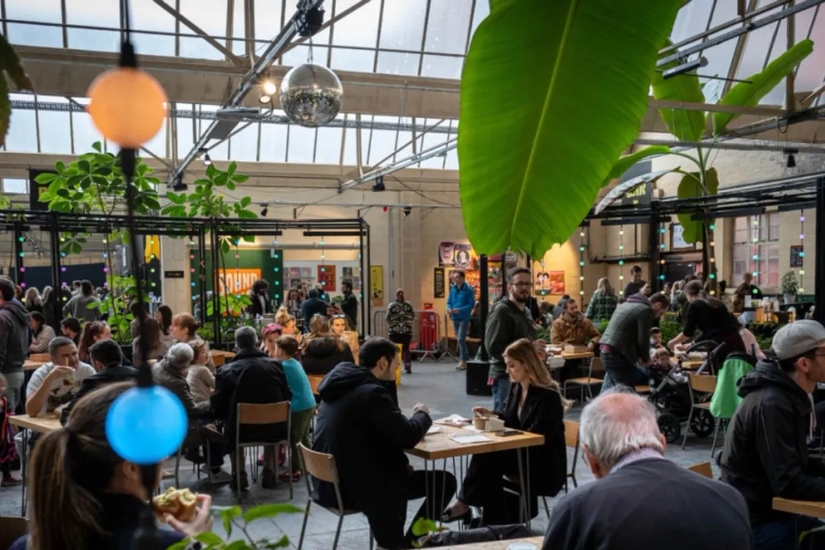 Busy indoor market or food hall with crowds of visitors seated at tables, colourful hanging lights, and lush green plants throughout a glass-roofed space.