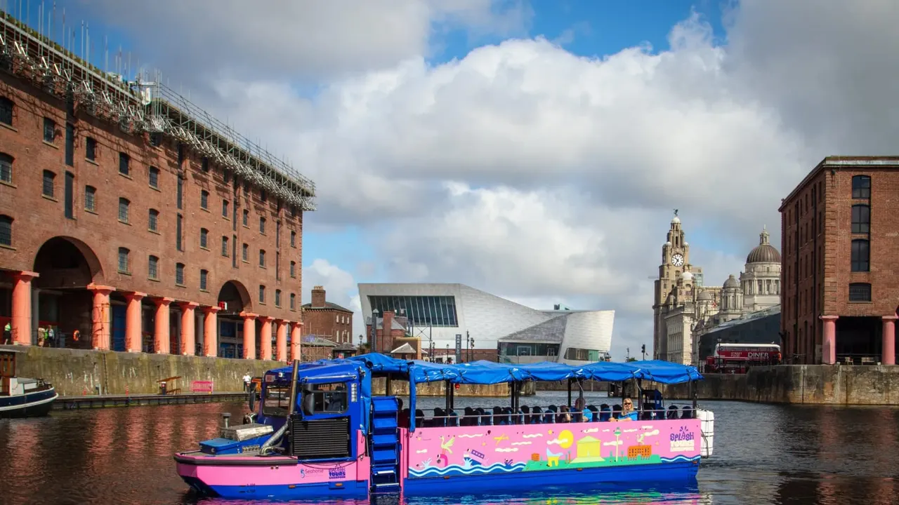 Colourful tour boat with blue canopy moored on the Albert Dock, with historic red-brick warehouses and Liverpool landmarks visible beyond.