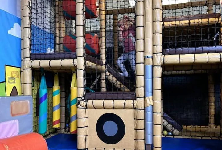 Child climbing a rope ladder inside a padded indoor play structure with tubular tunnels and netting.
