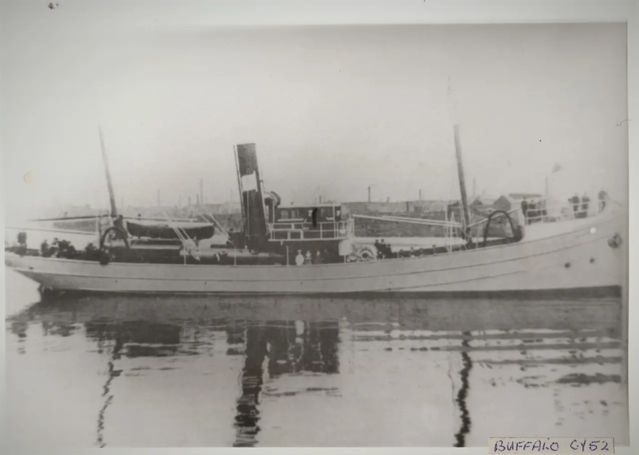 Vintage black and white photograph of a working fishing trawler moored in shallow water with industrial docks visible in the background.