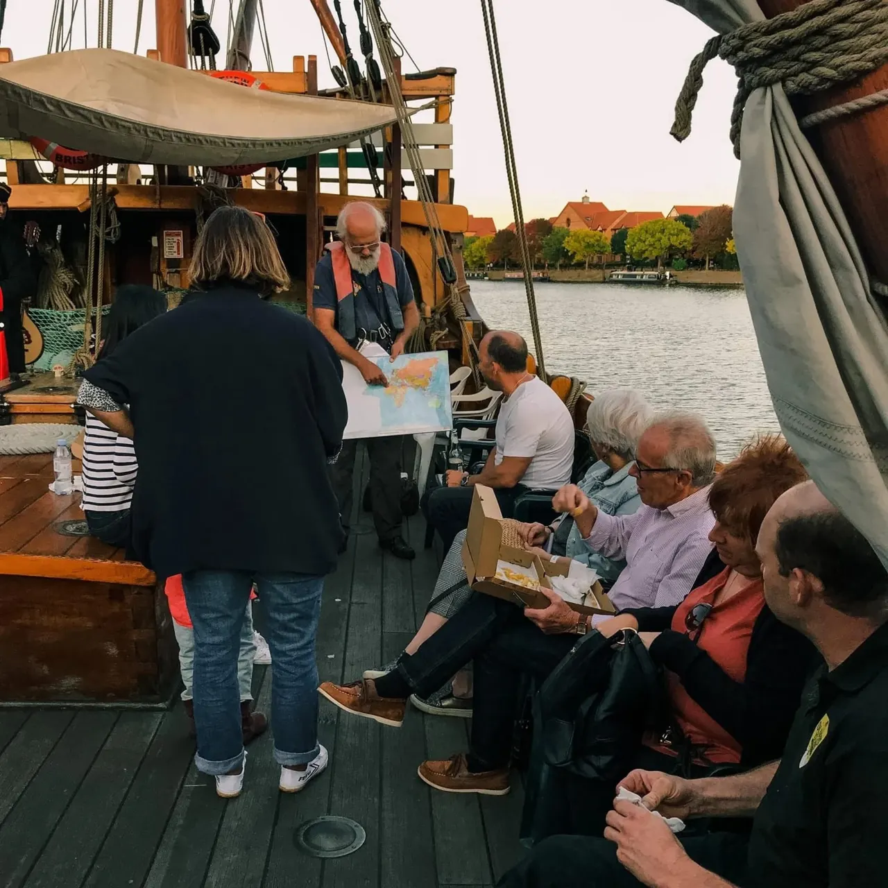 A guide holds a map whilst addressing passengers seated on the deck of a traditional wooden sailing vessel moored on a river.
