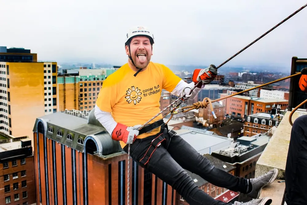 Man abseiling down the side of a tall building above a city skyline, wearing safety helmet and yellow charity shirt.