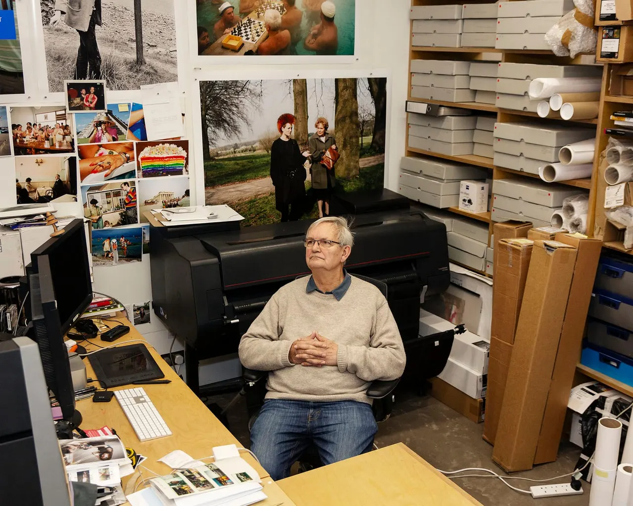 A man in glasses and a beige jumper sits in a cluttered creative studio surrounded by photographs, printing equipment, and storage shelves.