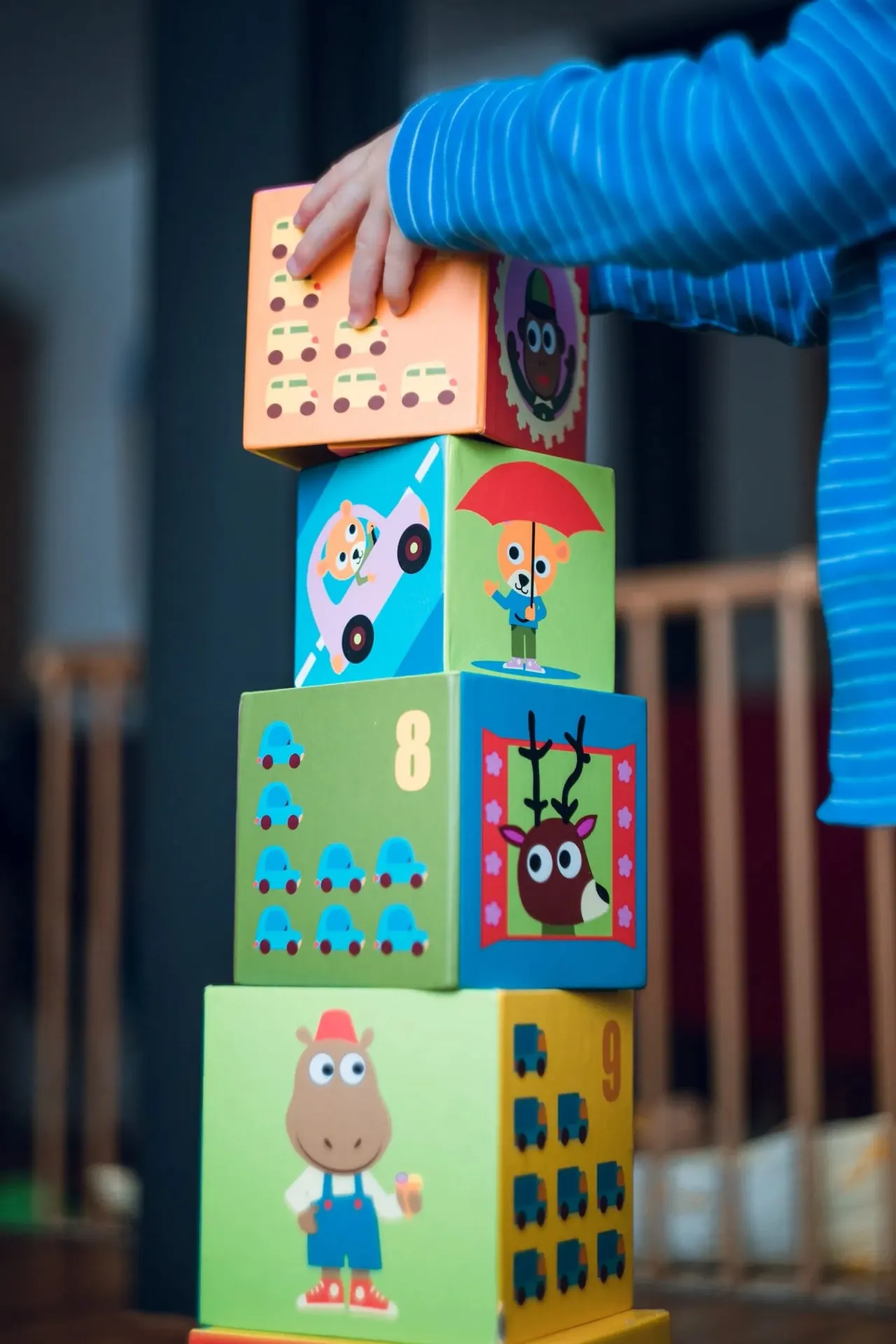 Child's hand touching a colourful stacked learning blocks tower with numbers, vehicles, and cartoon character illustrations.