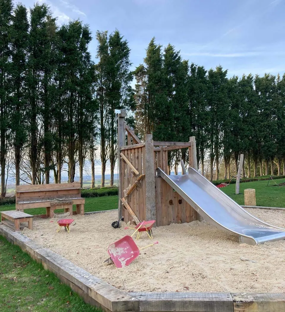 Children's play area with wooden climbing structure, slide and sand pit, surrounded by tall poplar trees.