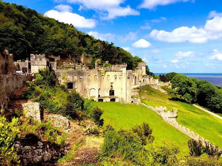 Medieval castle ruins on a hillside surrounded by lush green vegetation with sea visible in the distance.