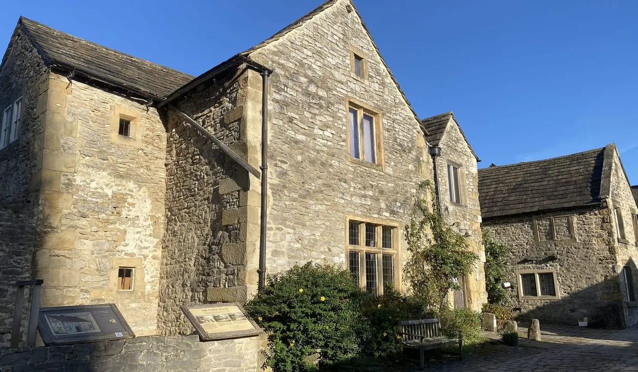 Stone buildings with gabled roofs under blue sky, information panels and bench visible in courtyard.