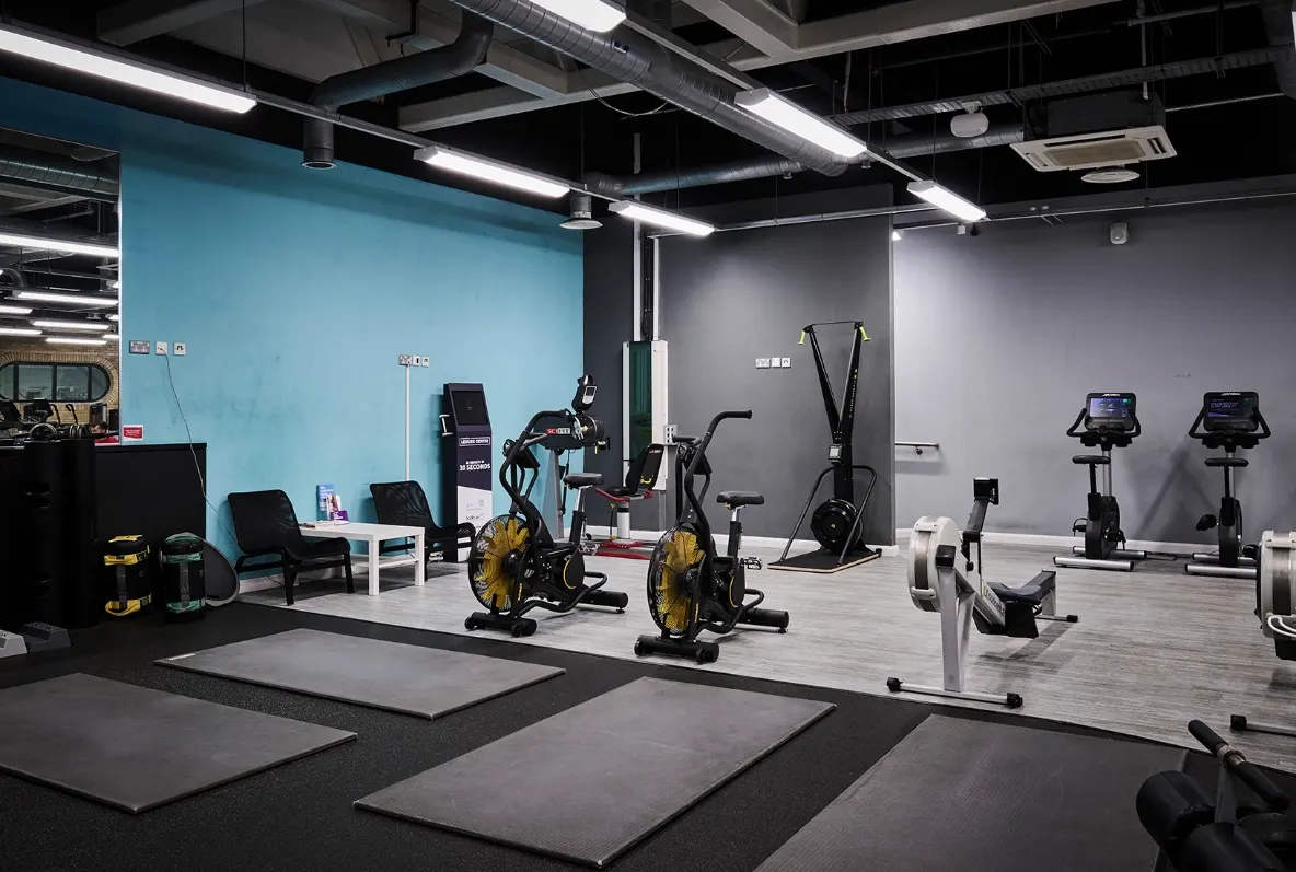 Modern gym interior with exercise bikes, stationary equipment, padded floor mats and blue and grey walls under bright lighting.