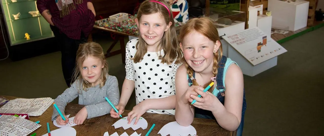 Three children smiling at a table whilst drawing or colouring with pencils during an indoor activity session.