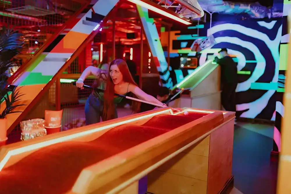 Woman playing mini golf on a neon-lit indoor course with colourful geometric designs and bright lighting.