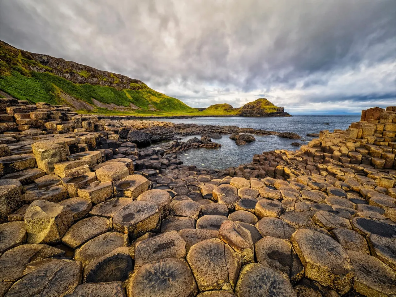 Hexagonal basalt columns on a rocky coastal shore with green hillside, small islands and cloudy sky beyond.