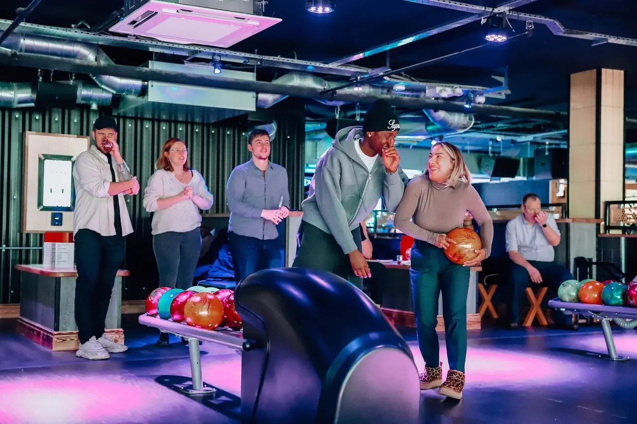 A group of people bowling together in a modern alley lit with pink and blue neon lighting.