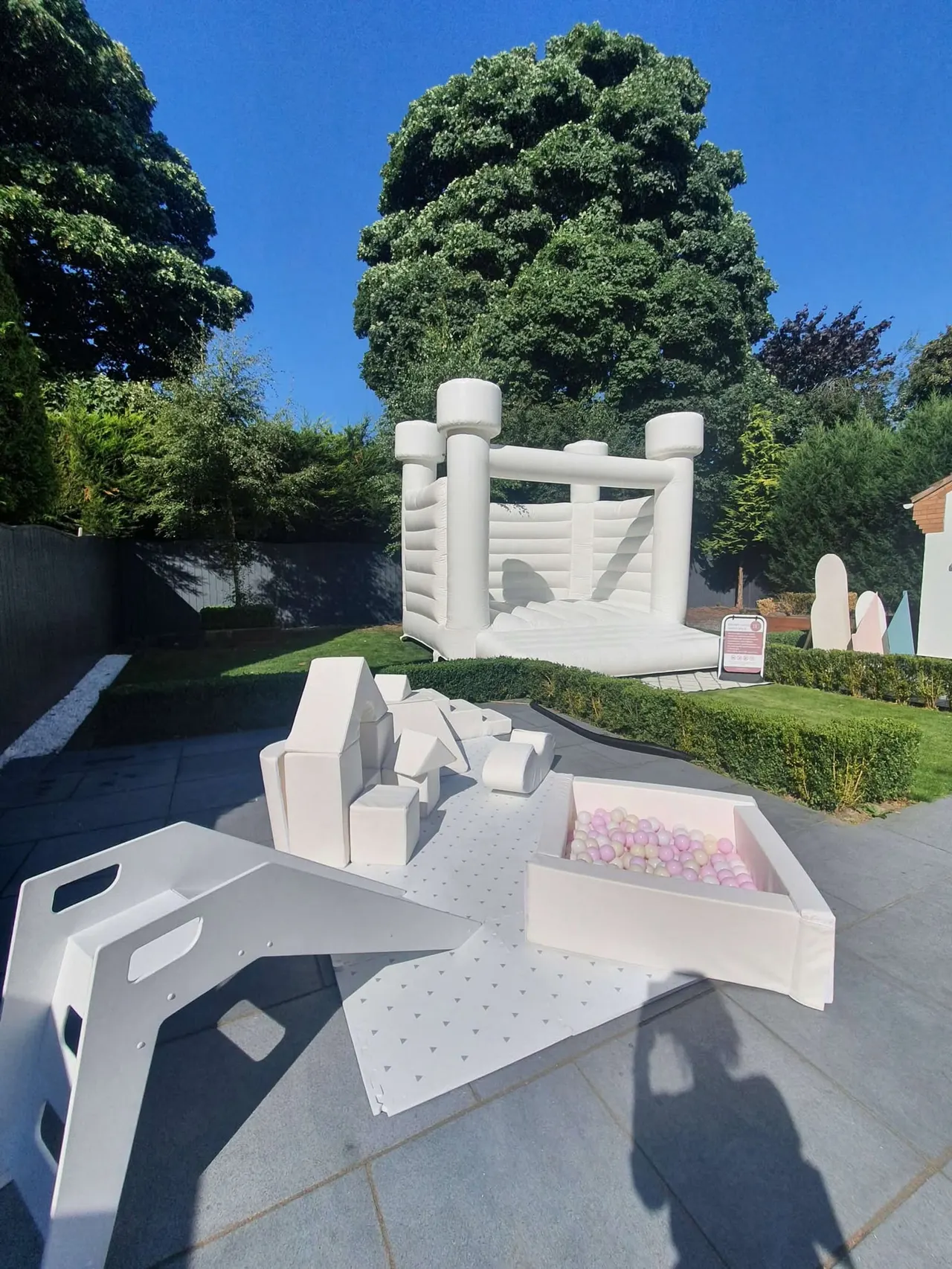 White inflatable bouncy castle with play equipment and pink ball pit in a garden setting on a sunny day.