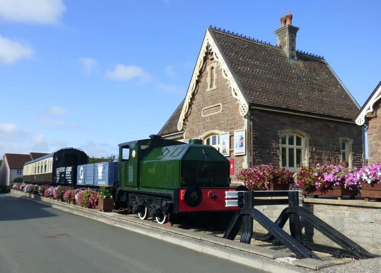 A green and red heritage railway locomotive and carriages parked on tracks beside a stone Victorian station building, with flowering baskets along the platform.