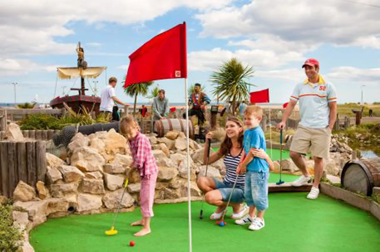 Families playing mini golf on a themed course with pirate ship scenery, red flags, and tropical plants under blue sky.