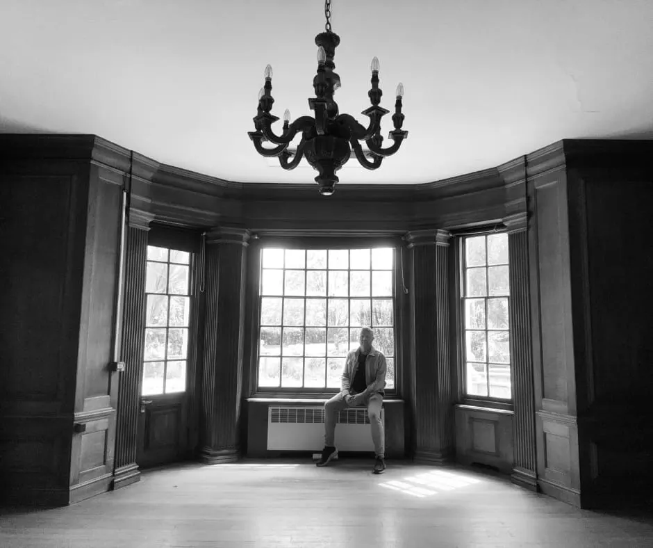Person sitting on window seat in elegant period room with black chandelier and bay windows.