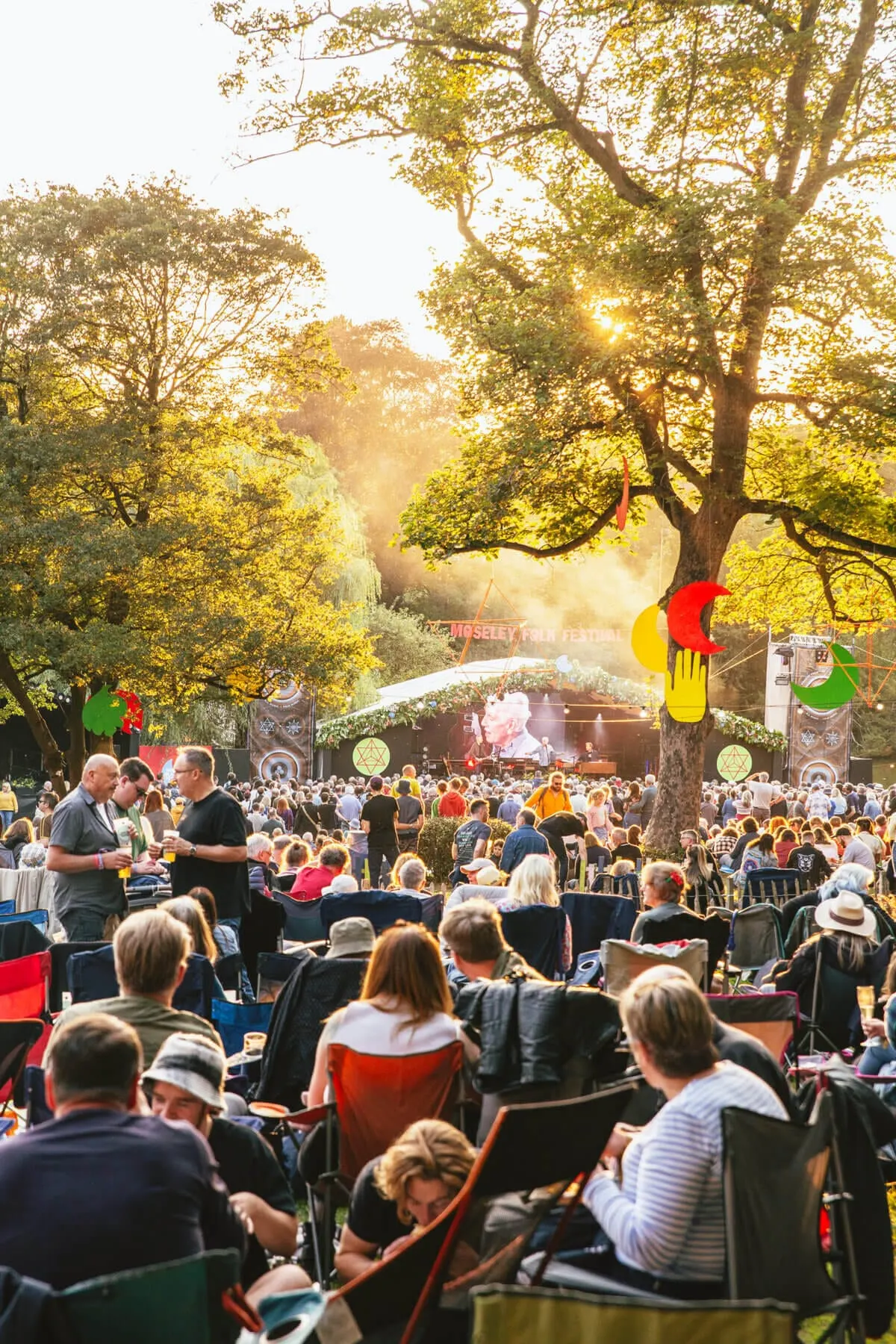 Outdoor festival crowd seated beneath tall trees with a stage and colourful decorations in the distance during golden-hour sunshine.