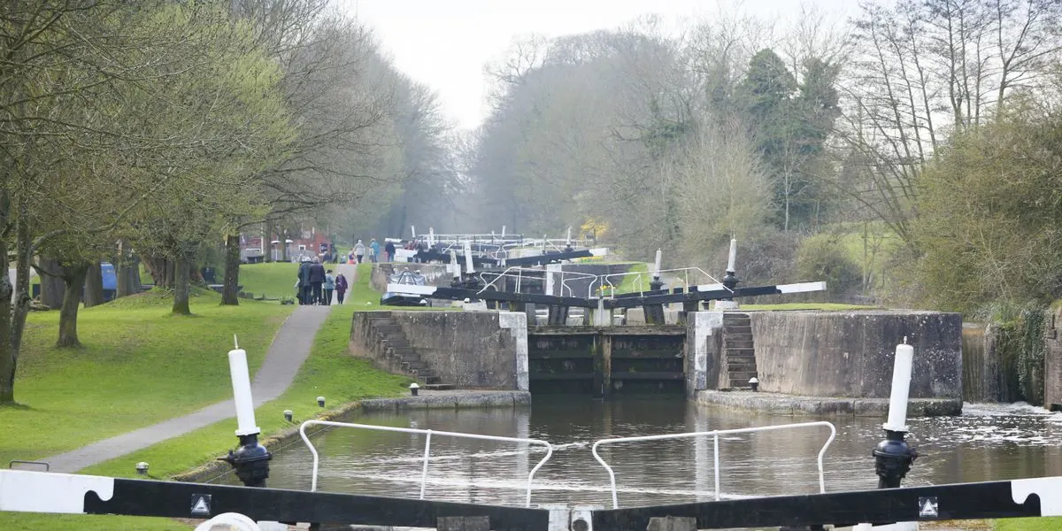 Narrowboats moored on a tree-lined canal with visitors walking along the towpath and lock gates in the distance.
