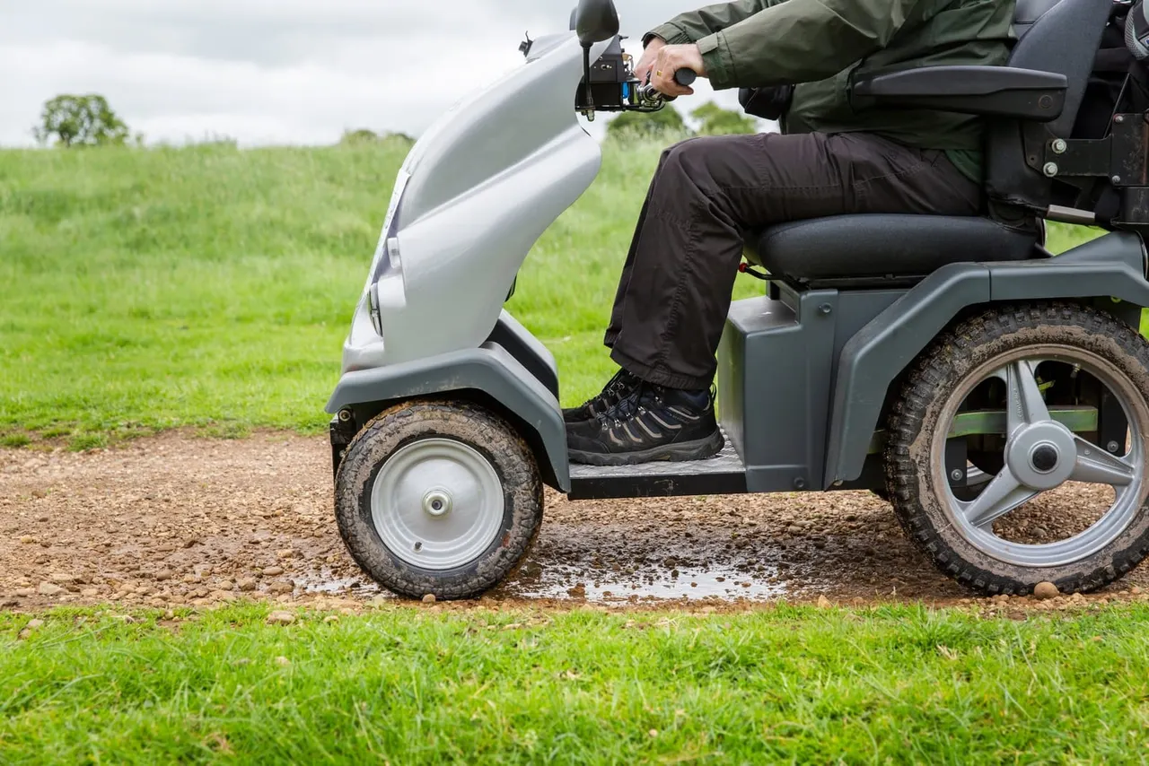 Person operating a mobility scooter on a gravel path beside green fields.