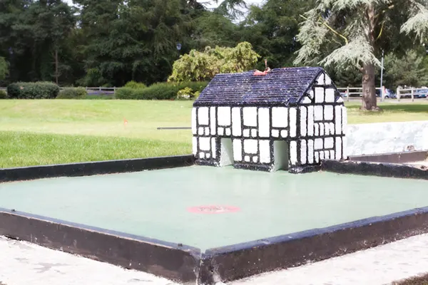A miniature Tudor-style building structure on a pale green mini golf course surrounded by trees and parkland.