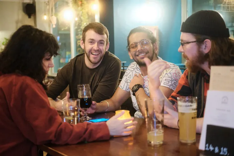 A group of five people gathered around a table with drinks, smiling and chatting together in a warmly lit indoor venue.