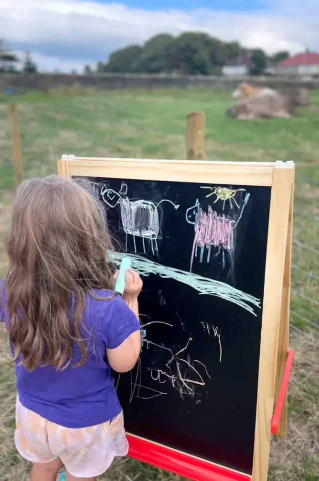 Young child drawing with chalk on a blackboard in a rural farm setting with fields and trees behind.