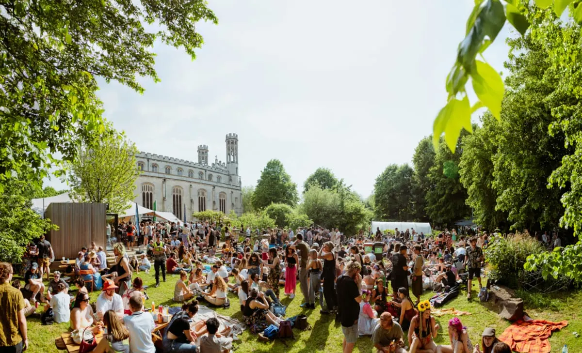 Large outdoor gathering with hundreds of visitors picnicking and socialising on a grassy park area framed by mature trees, with a historic stone building featuring twin towers in the background.