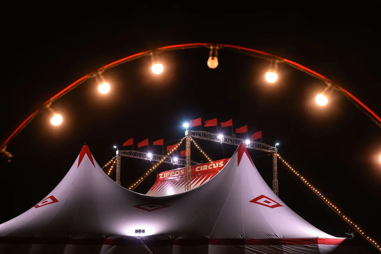 Illuminated circus big top tent with red and white striped structure, ornate arched entrance lit by warm bulbs, and decorated framework above at night.